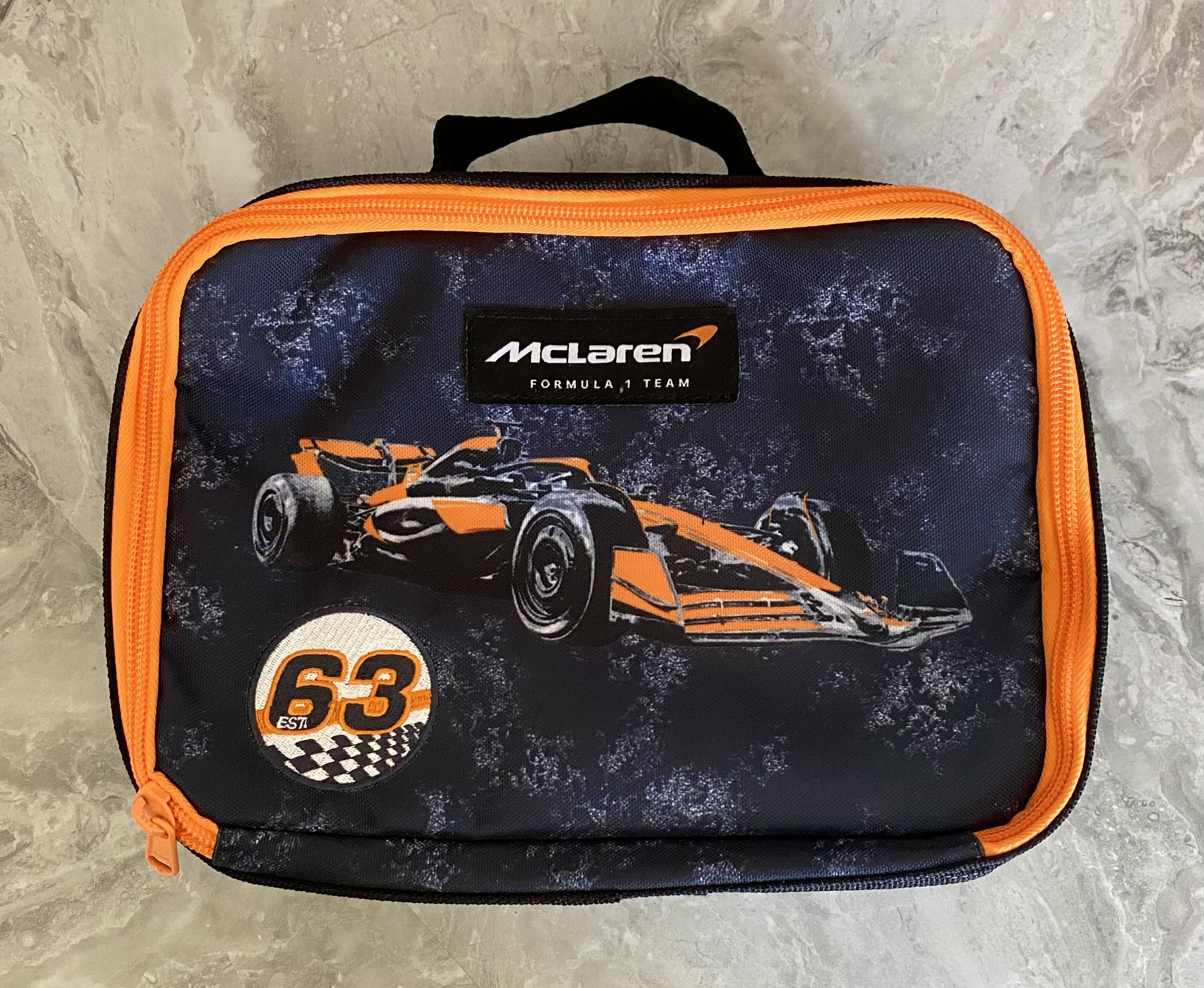 A rectangular McLaren Formula 1 Team lunch bag with a black and orange color scheme. It features a stylized orange and black Formula 1 car on the front, a stitched Est. 63 patch with a checkered flag motif, and bright orange piping along the zipper and edges.