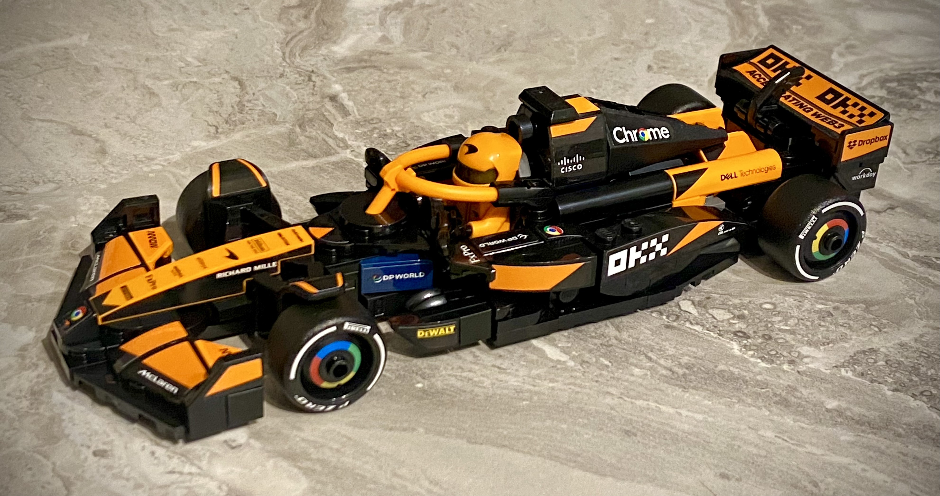 A LEGO McLaren Formula 1 car in black and papaya orange with detailed sponsor logos including Chrome, Cisco, Dell Technologies, and Dropbox. The car has a halo device, a miniature driver figure in a matching helmet, and hard-compound Pirelli tires.