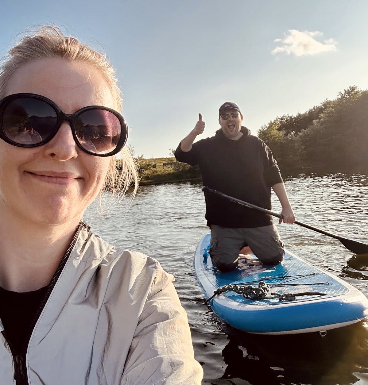 Two people are enjoying a sunny day on the water. A woman in large round sunglasses and a light jacket smiles at the camera in the foreground. Behind her, a man kneels on a blue paddleboard, giving a thumbs-up with one hand and holding a paddle with the other.