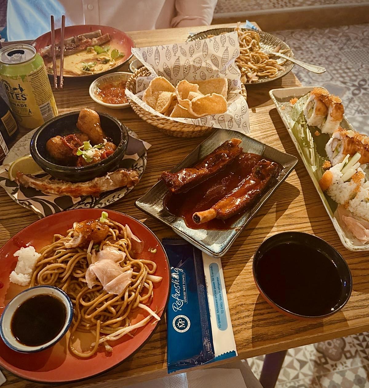 Table filled with various Asian-inspired dishes including noodles, sushi rolls, barbecue ribs, prawn crackers, spring rolls, and dipping sauces. A man in a pink shirt sits at the top of the frame.