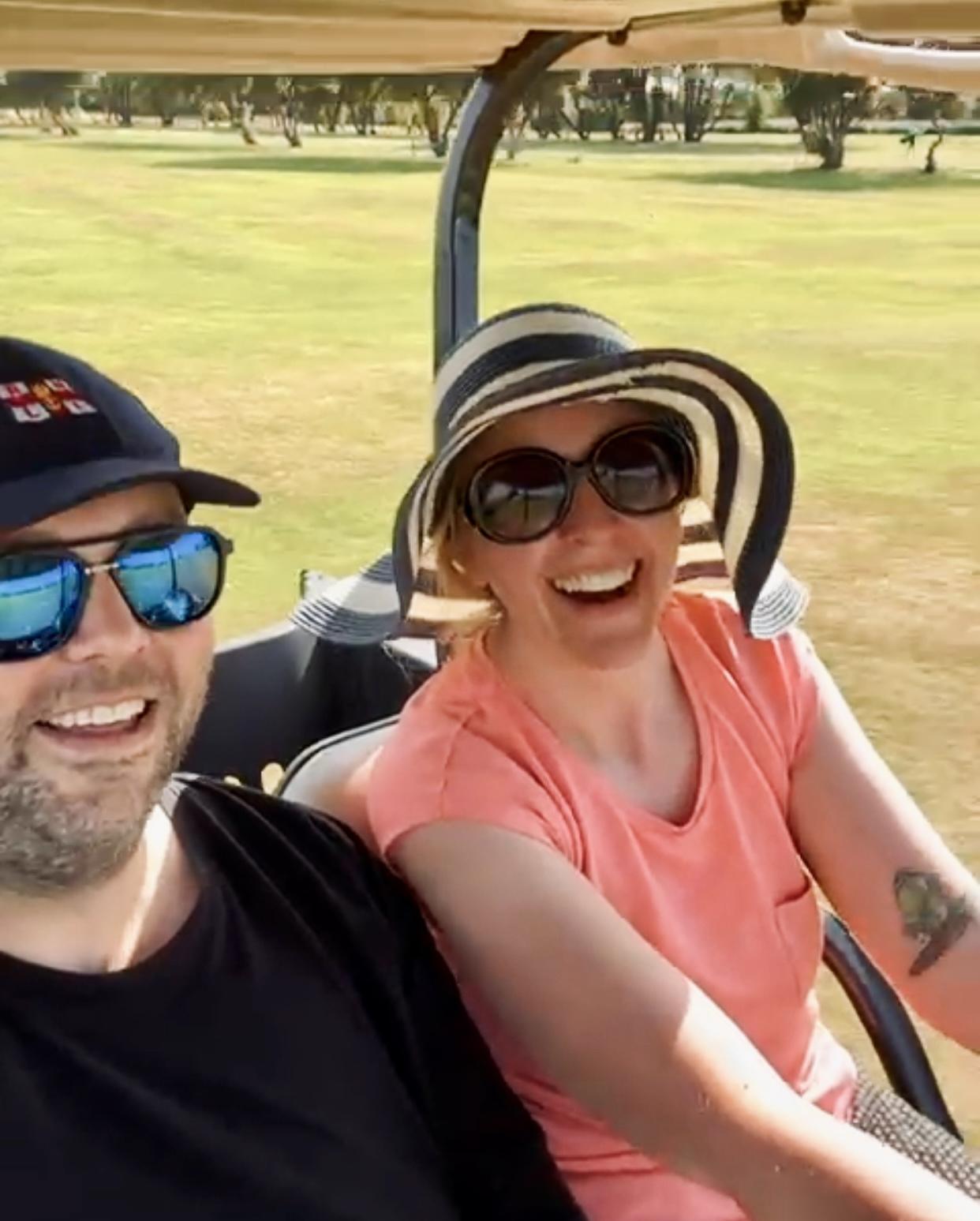 Two people are sitting in a golf buggy. The person on the left wears a black shirt, sunglasses with blue lenses, and a dark blue cap. The person on the right wears a striped sun hat, large sunglasses, and a coral short-sleeve top. Both are smiling.