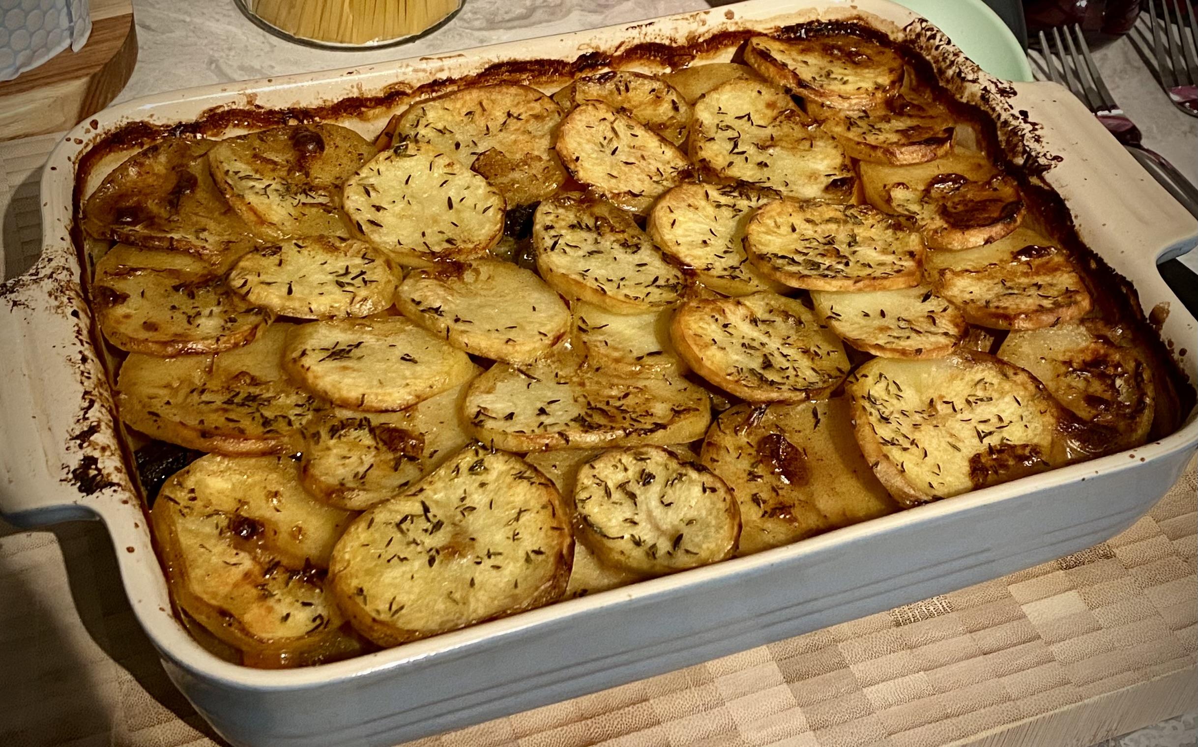 A rectangular oven dish filled with a layered potato bake. Thin potato slices are browned and crisp on top, sprinkled with herbs, and arranged neatly in overlapping rows. The edges of the dish are slightly browned from baking.