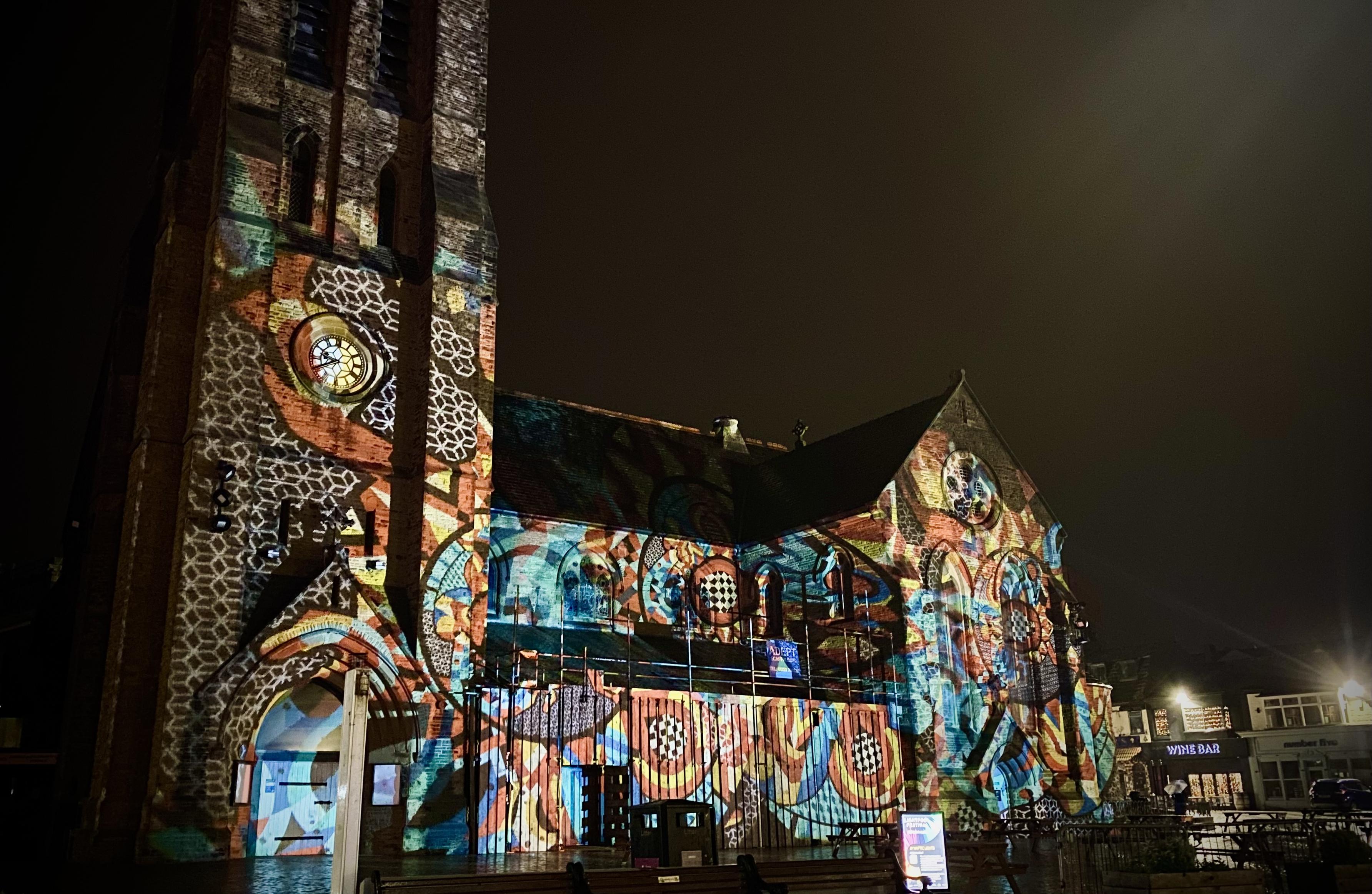Church building covered in colourful projected light art featuring geometric and abstract designs during a night-time display.