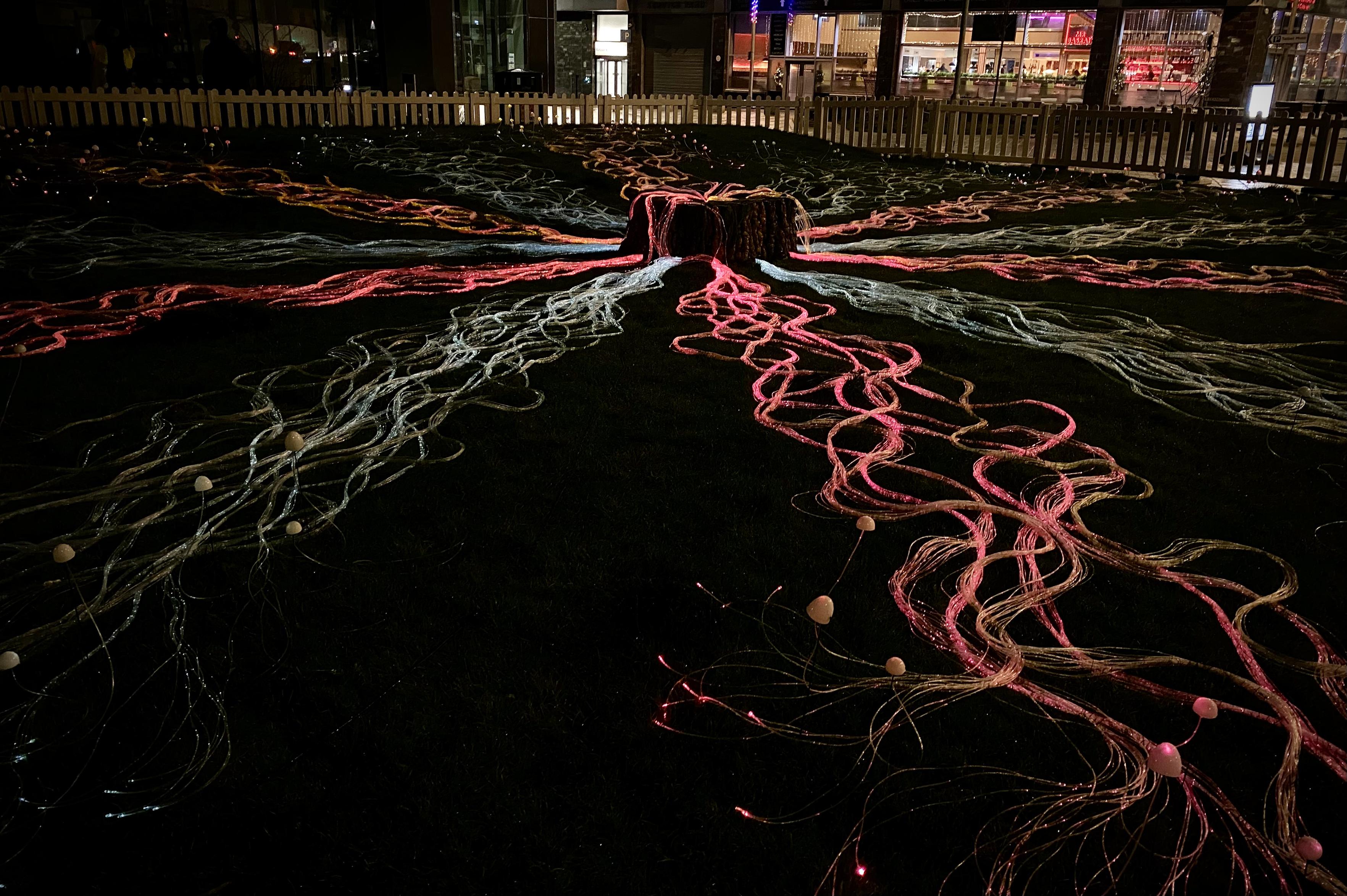 Tree stump installation surrounded by glowing fibre-optic strands spreading outward like mycelium (the root-like network of a fungus, made of a mass of branching, thread-like strands called hyphae), lit in red and white in dark surroundings.