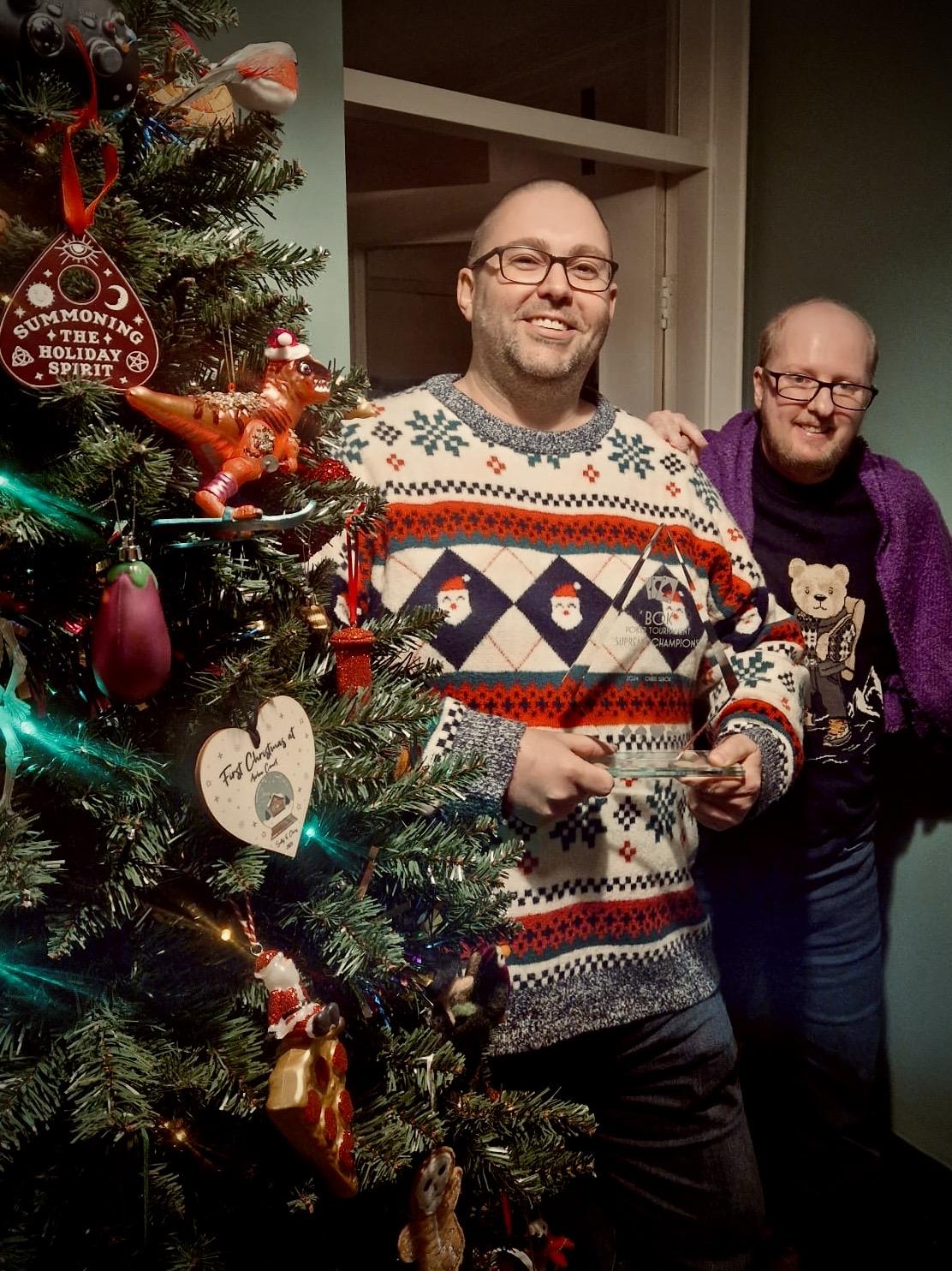 Two men stand beside a decorated Christmas tree. Both wear patterned jumpers, one with a Santa design and the other with a bear. One man holds a small trophy while the other rests a hand on his shoulder.